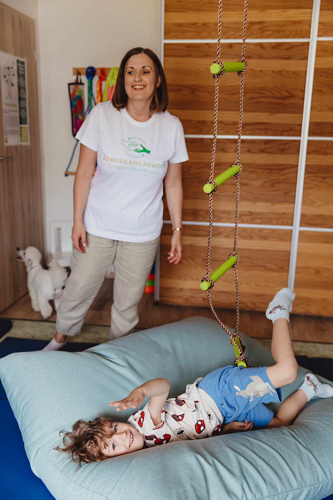 Usmievavá terapeutka stojí pri dieťati na podložke, v pozadí drevená stena a zelené terapeutický rebrík. Senzorická integrácia